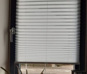 Foto eines Fensters mit geschlossenen grauen Jalousien. Die Lamellen der Jalousien blockieren das Tageslicht, lassen jedoch leichte Schatten und Konturen erkennen. Das Bild ist von einem blauen Rahmen mit Messmarkierungen an den Ecken umgeben.