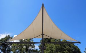 Beige Sonnensegel, gespannt über einem Metallgestänge, bietet Schatten an einem sonnigen Tag, vor dem Hintergrund von blauem Himmel und grünen Bäumen.
