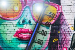 Buntes Graffiti einer Frau mit übergroßen Sonnenbrillen auf einer Backsteinmauer, hinterlegt von einem großen Thermometer, das hohe Temperaturen anzeigt.